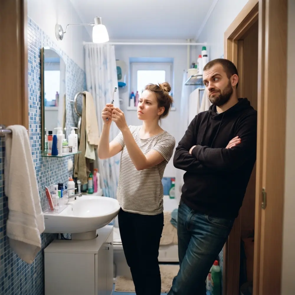 Couple in a small bathroom — one holds up a toothbrush, the other leans on the doorframe, unconvinced